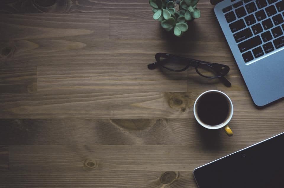 A wooden desk with a keyboard, a cup of coffee and a pair of glasses