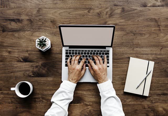 Hands typing on a laptop at a wooden desk beside a coffee cup