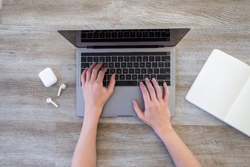 Hands typing on a laptop beside wireless earbuds and a notebook