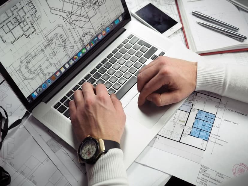 Hands typing on a laptop surrounded by printed plans and pens