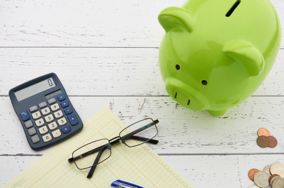 A green piggy bank beside a calculator, glasses and a notepad
