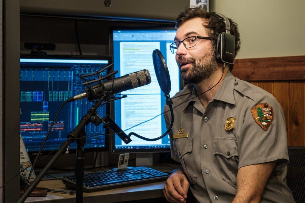 A person speaking into a studio microphone in front of computer monitors