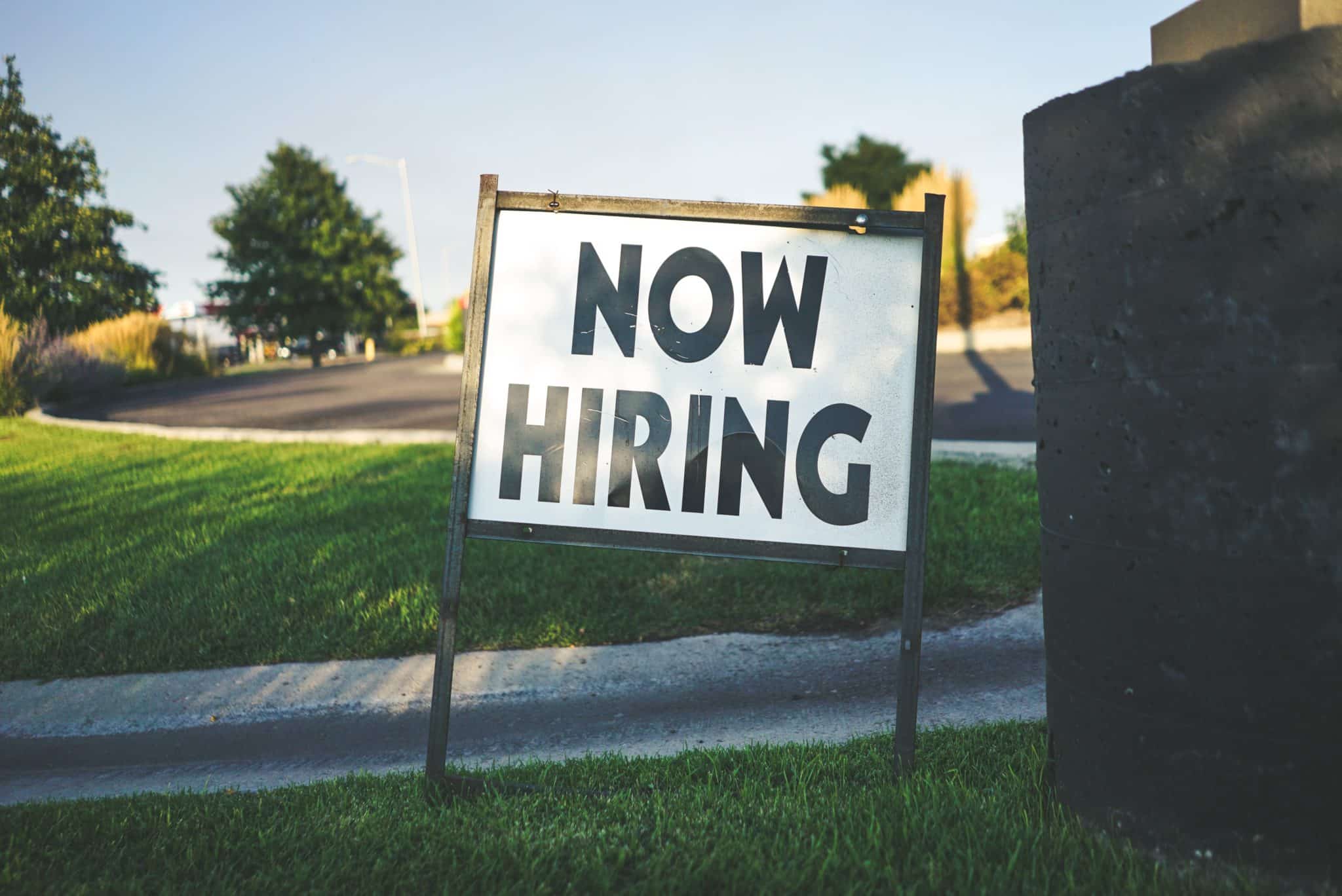 A now hiring sign staked in the grass beside a path